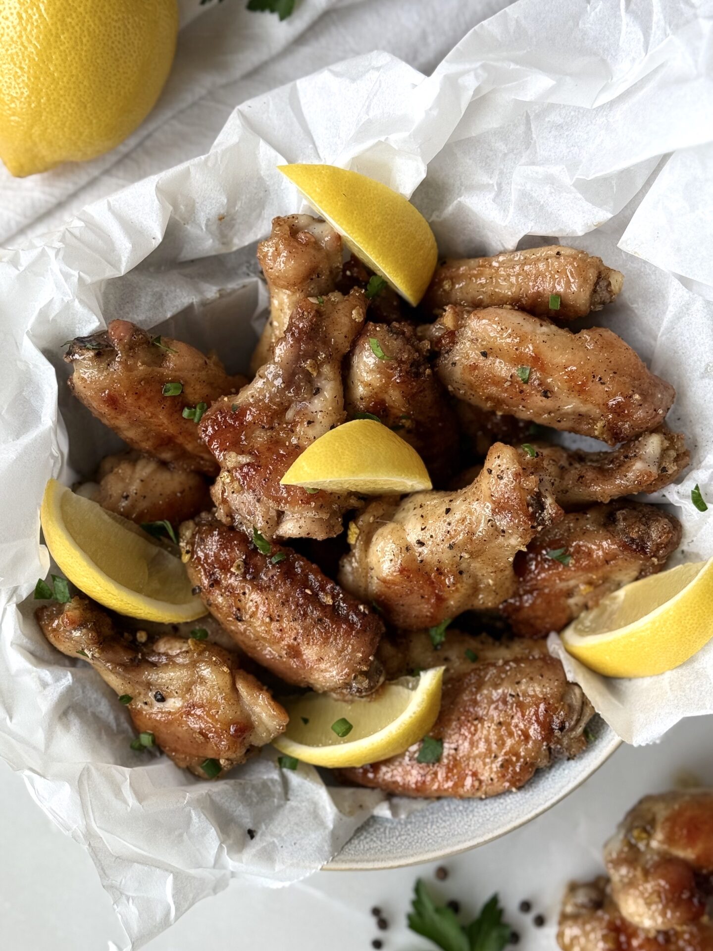 A bowl of golden Lemon Pepper Chicken Wings is seen from above, garnished with lemon wedges, parsley and chives