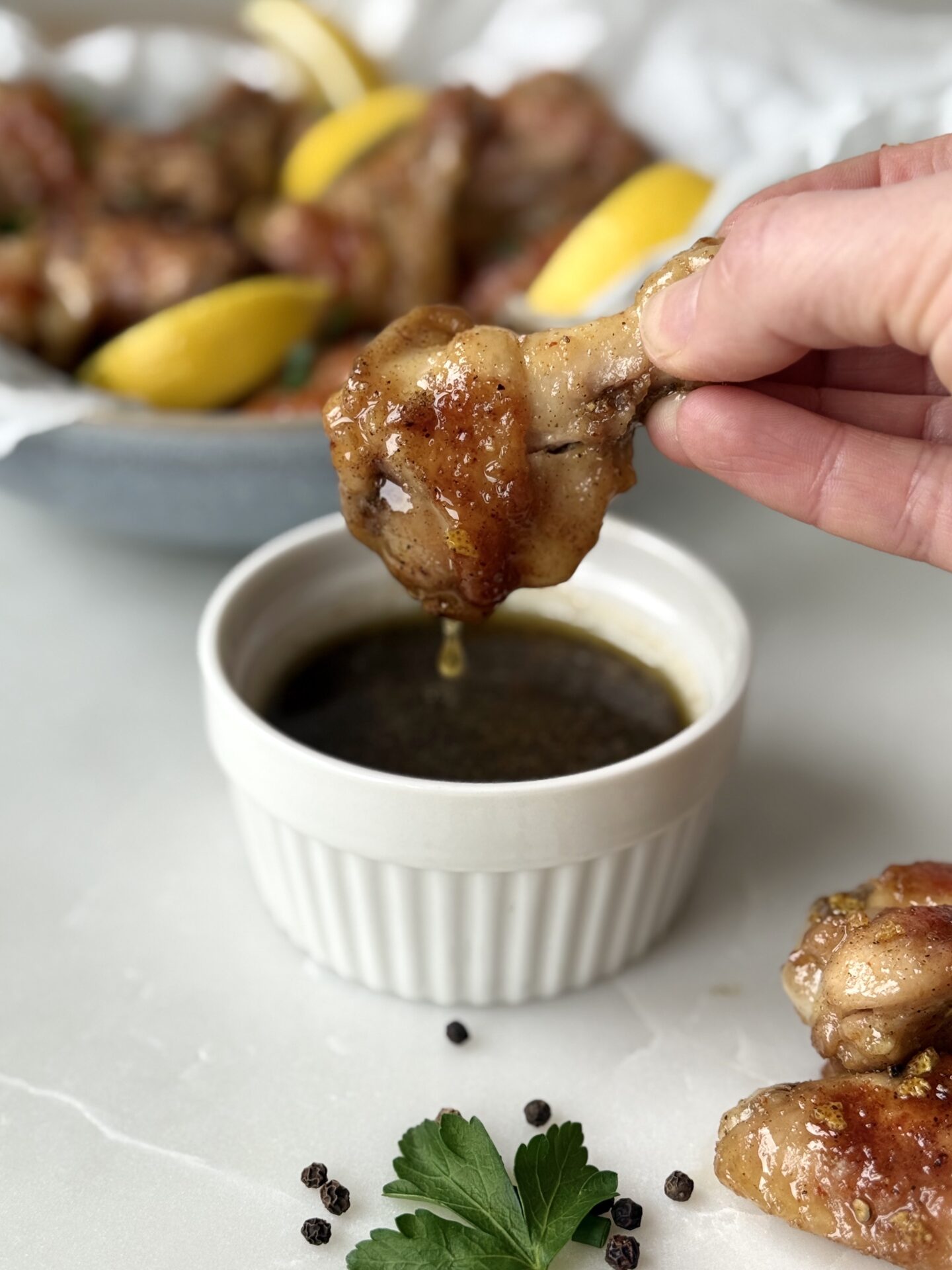 A hand is seen dipping a lemon pepper chicken wing into a bowl of sauce.  A platter of wings and lemon wedges is seen in the background.