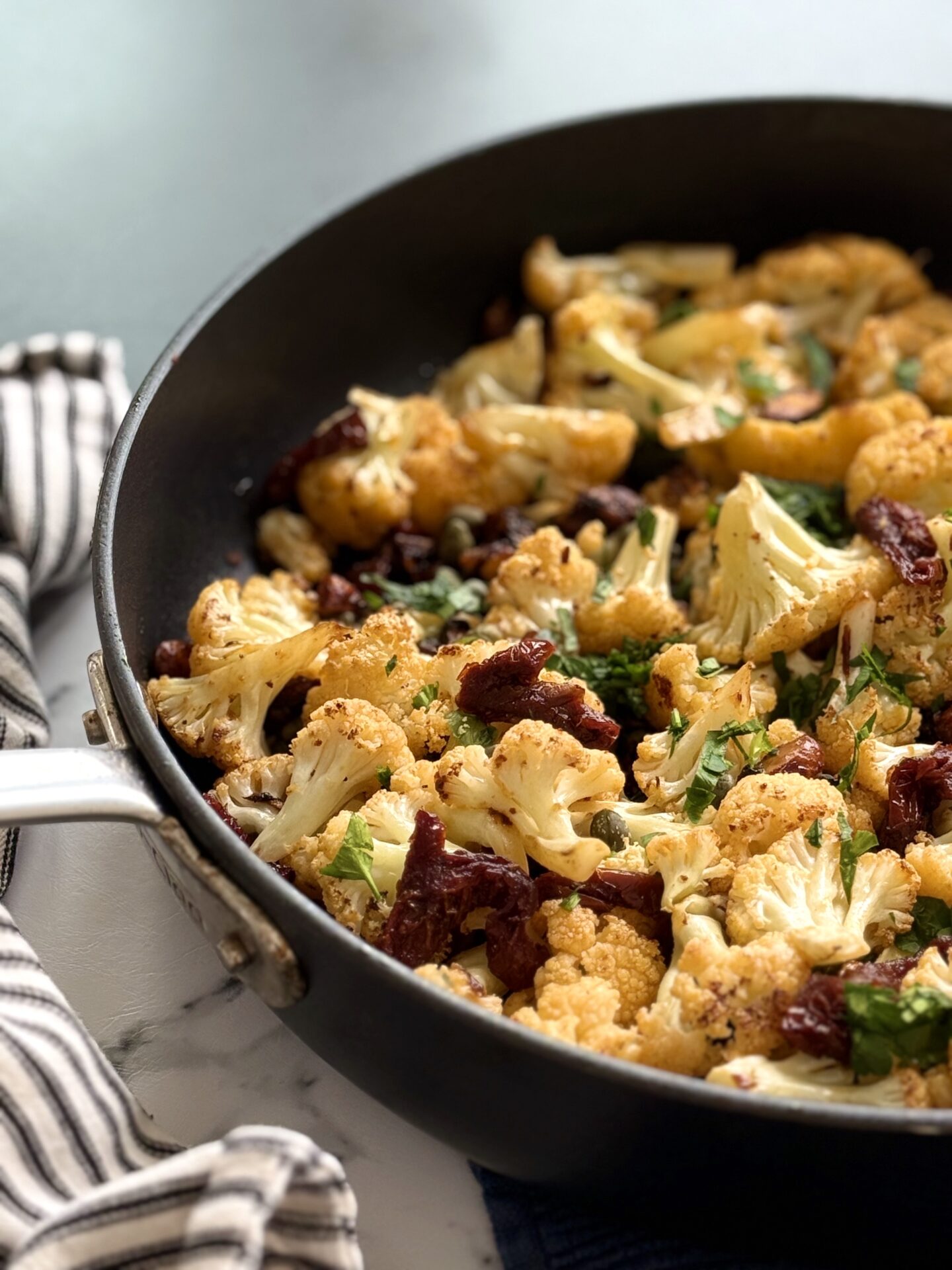 A large black skillet full of Roasted Cauliflower with Sundried Tomatoes, Capers and Crunchy Almonds is seen up close with a black and white striped towel wrapped around the skillet handle.