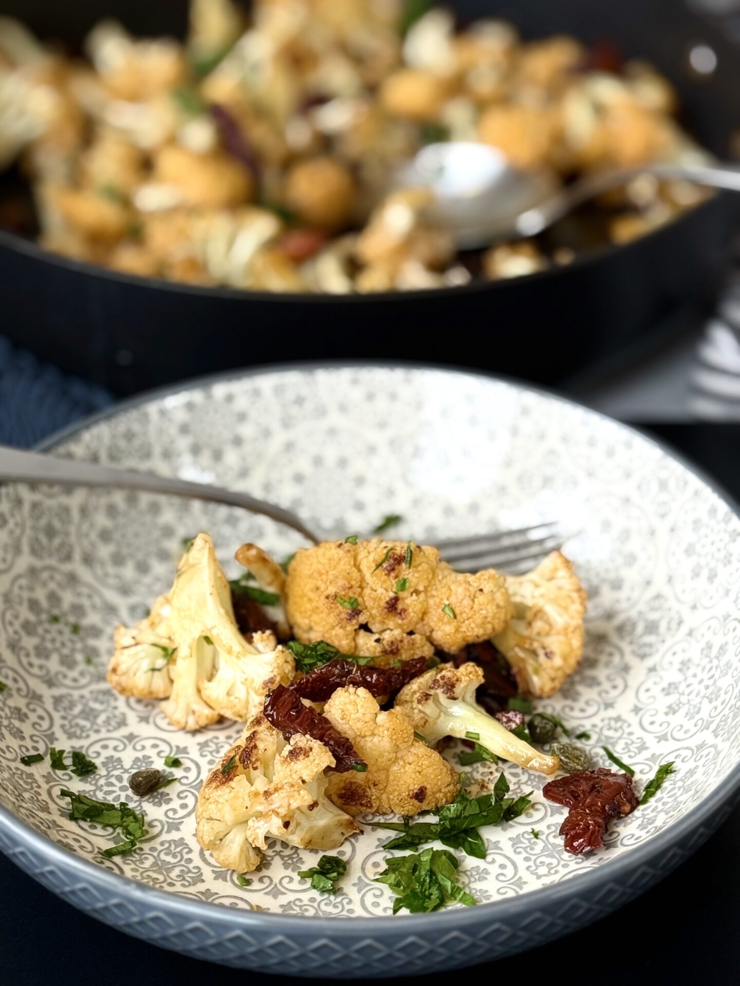 A serving of Roasted Cauliflower with Sundried Tomatoes, Capers and Crunchy Almonds is seen in the foreground in a grey patterned bowl.  In the background a black skillet full of this cauliflower dish can be seen.