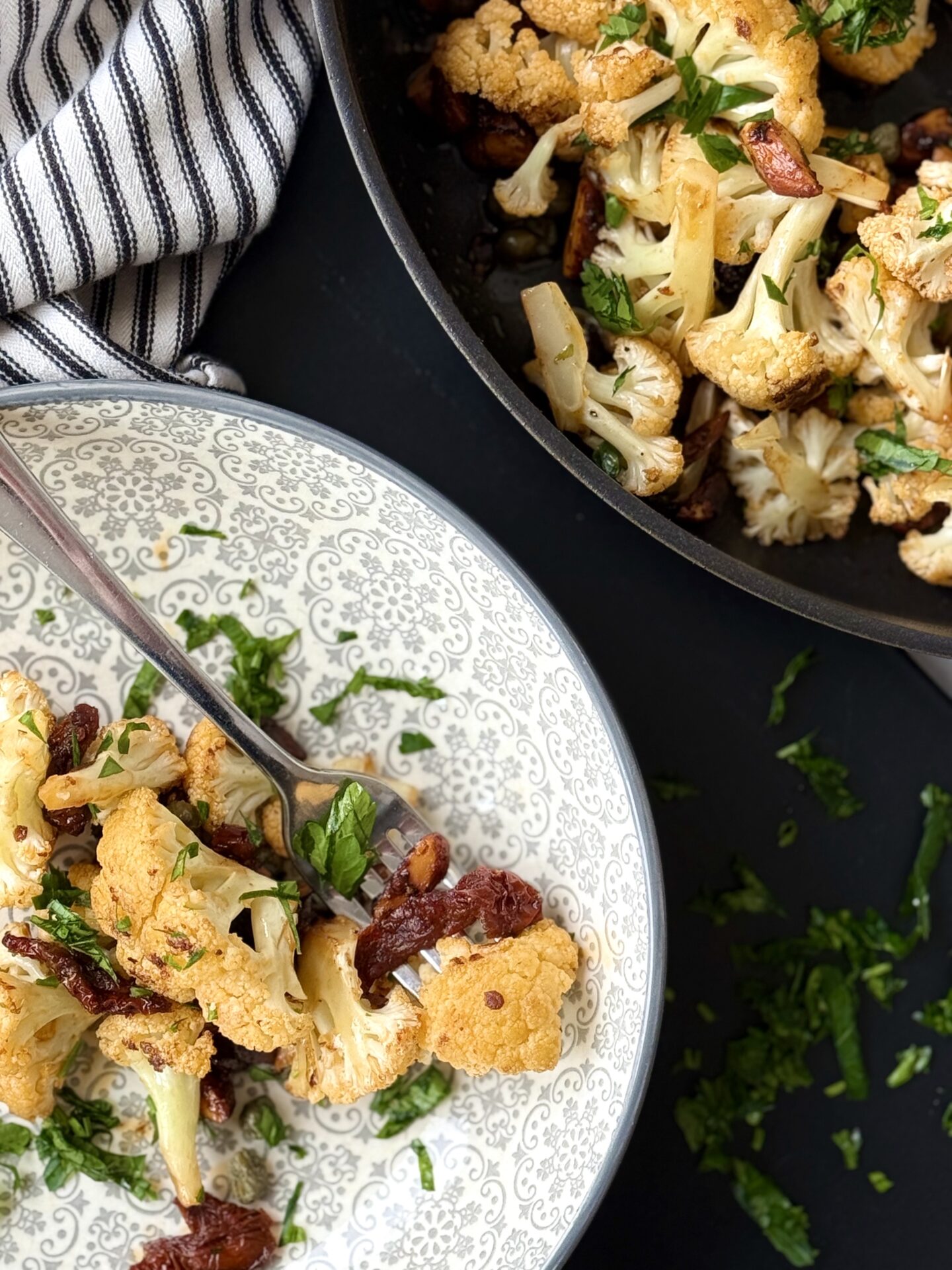 In the lower left hand corner, a grey patterned bowl is seen with a serving of Roasted Cauliflower with Sundried Tomatoes, Capers and Crunchy Almonds.  In the opposite corner the black skillet containing the cauliflower dish can be seen.