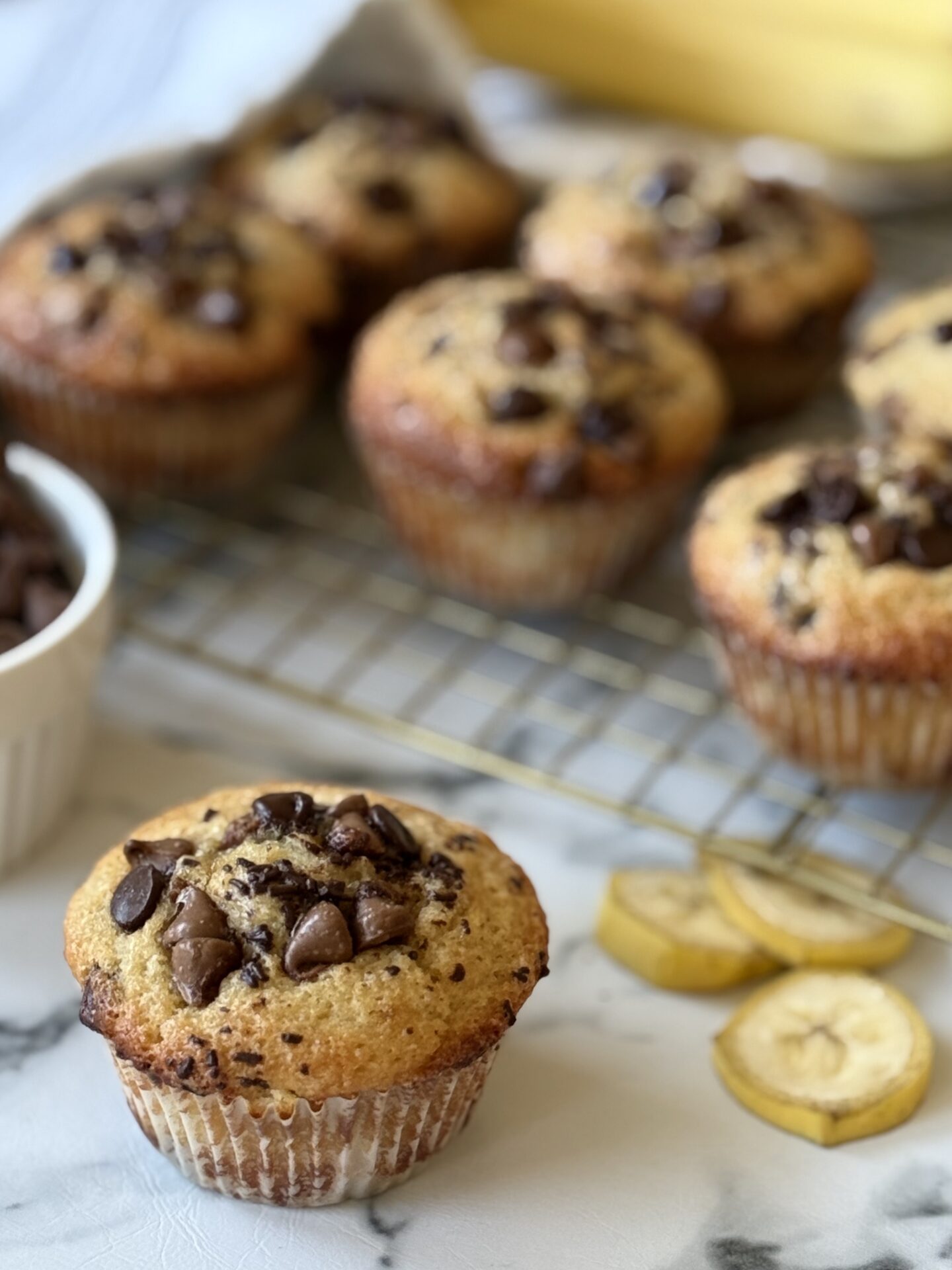 A Sour Cream Banana Chocolate Chip Muffin is seen in the foreground, with other muffins, banana slices, whole bananas and chocolate chips seen in the background.