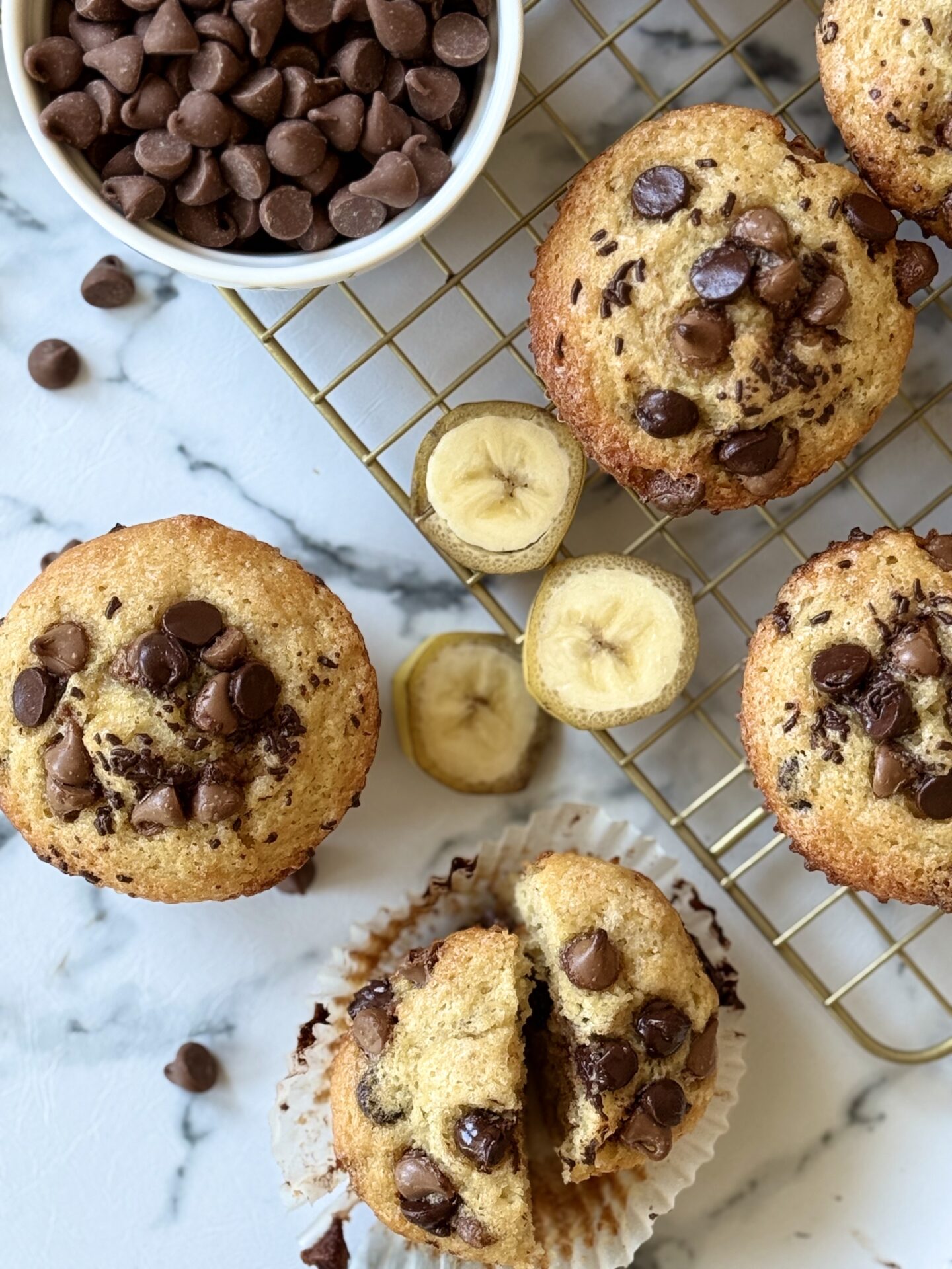 Four Sour Cream Banana Chocolate Chip Muffins are seen from above, surrounded by slices of fresh bananas and chocolate chips.  