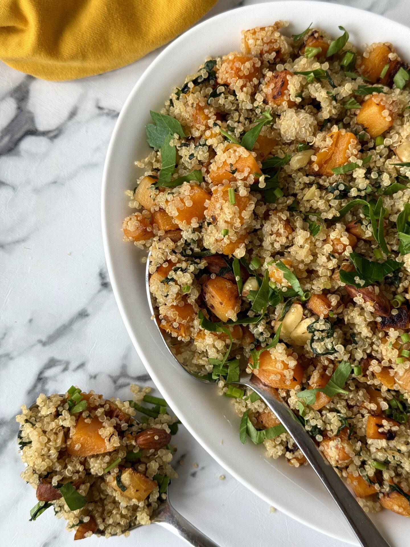 Half of a serving platter full of quinoa salad with butternut squash, spinach and crunchy almonds is seen on white marble counter.  A serving spoon overflowing with the salad sits beside the platter.