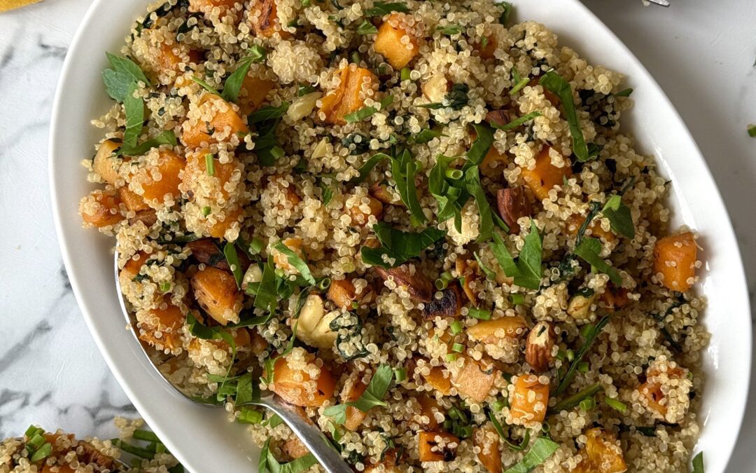 A platter of quinoa salad with butternut squash, spinach and crunchy almonds is seen from above on a white marble counter, surrounded by serving utensils and a yellow dinner napkin.