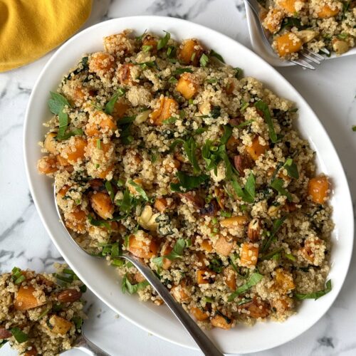 A platter of quinoa salad with butternut squash, spinach and crunchy almonds is seen from above on a white marble counter, surrounded by serving utensils and a yellow dinner napkin.