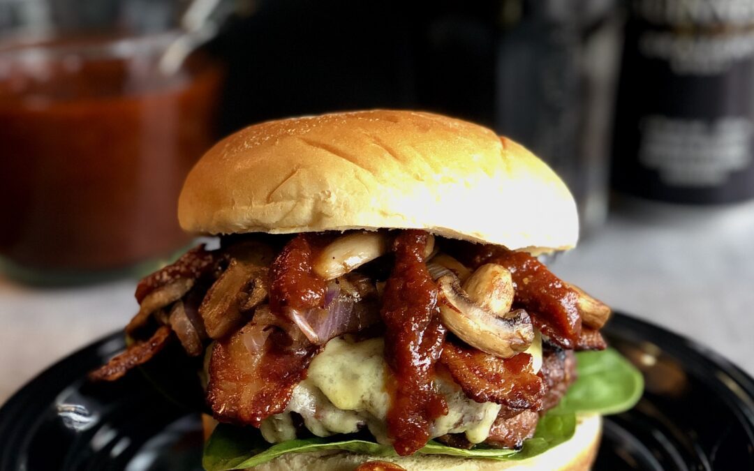 A Guinness Bacon Burger with Irish Cheddar and Mushrooms is seen in the foreground, dripping with homemade Guinness barbecue sauce. A can and pint of Guinness beer are seen in the background.