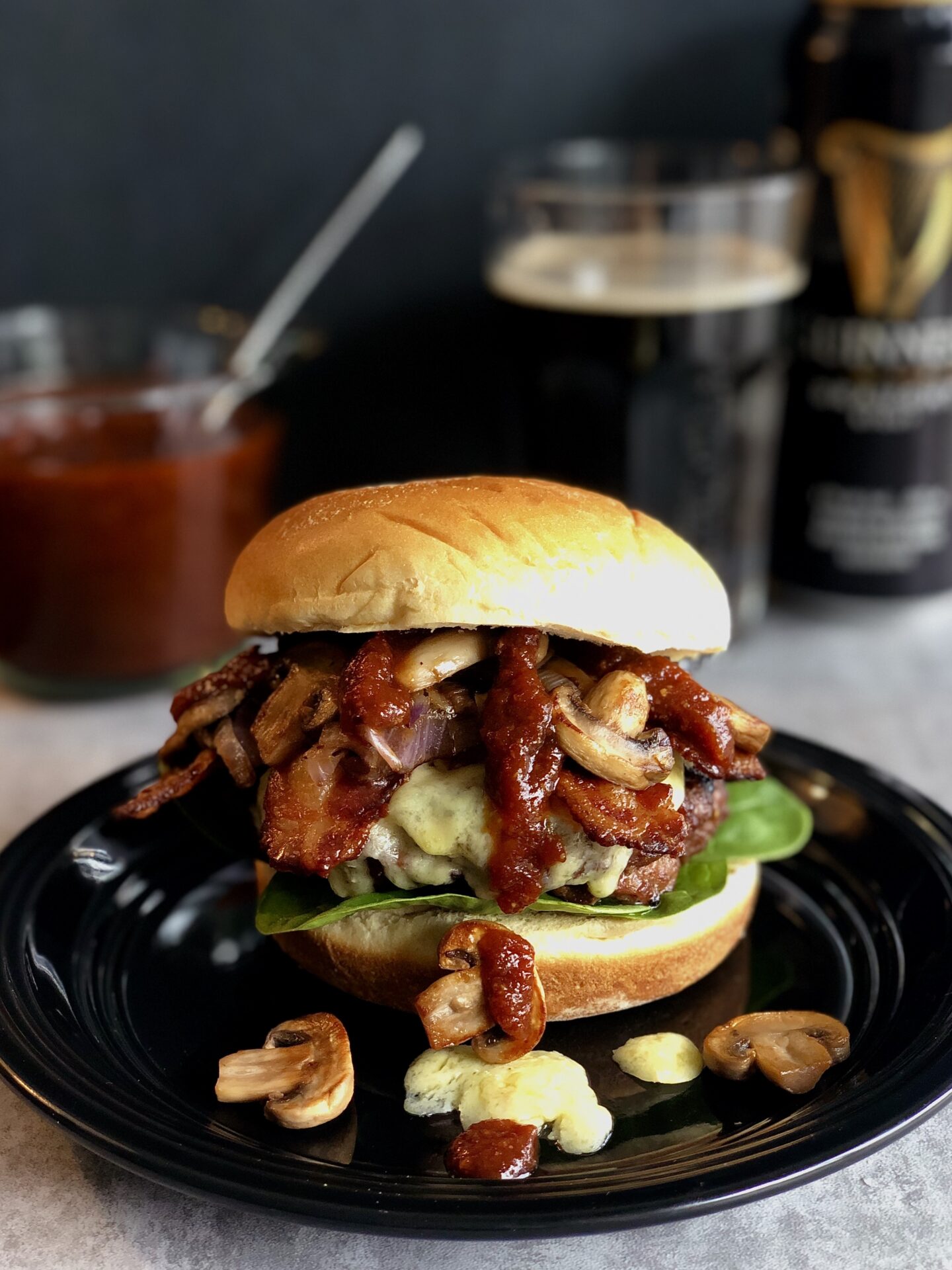 A Guinness Bacon Burger with Mushrooms and Irish Cheddar is seen on a black plate. Guinness Barbecue sauce and a can of Guinness beer can be seen in the background