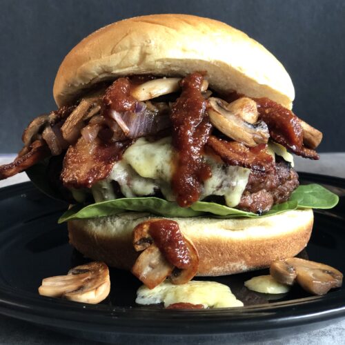 A Guinness Bacon Burger with Irish Cheddar and Mushrooms is seen close up. Guinness barbecue sauce drips down and golden sautéed mushrooms tumble out of the burger onto the black plate underneath.