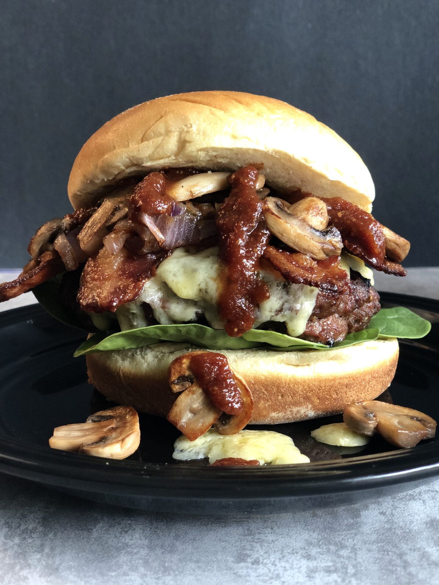 A Guinness Bacon Burger with Irish Cheddar and Mushrooms is seen close up. Guinness barbecue sauce drips down and golden sautéed mushrooms tumble out of the burger onto the black plate underneath.