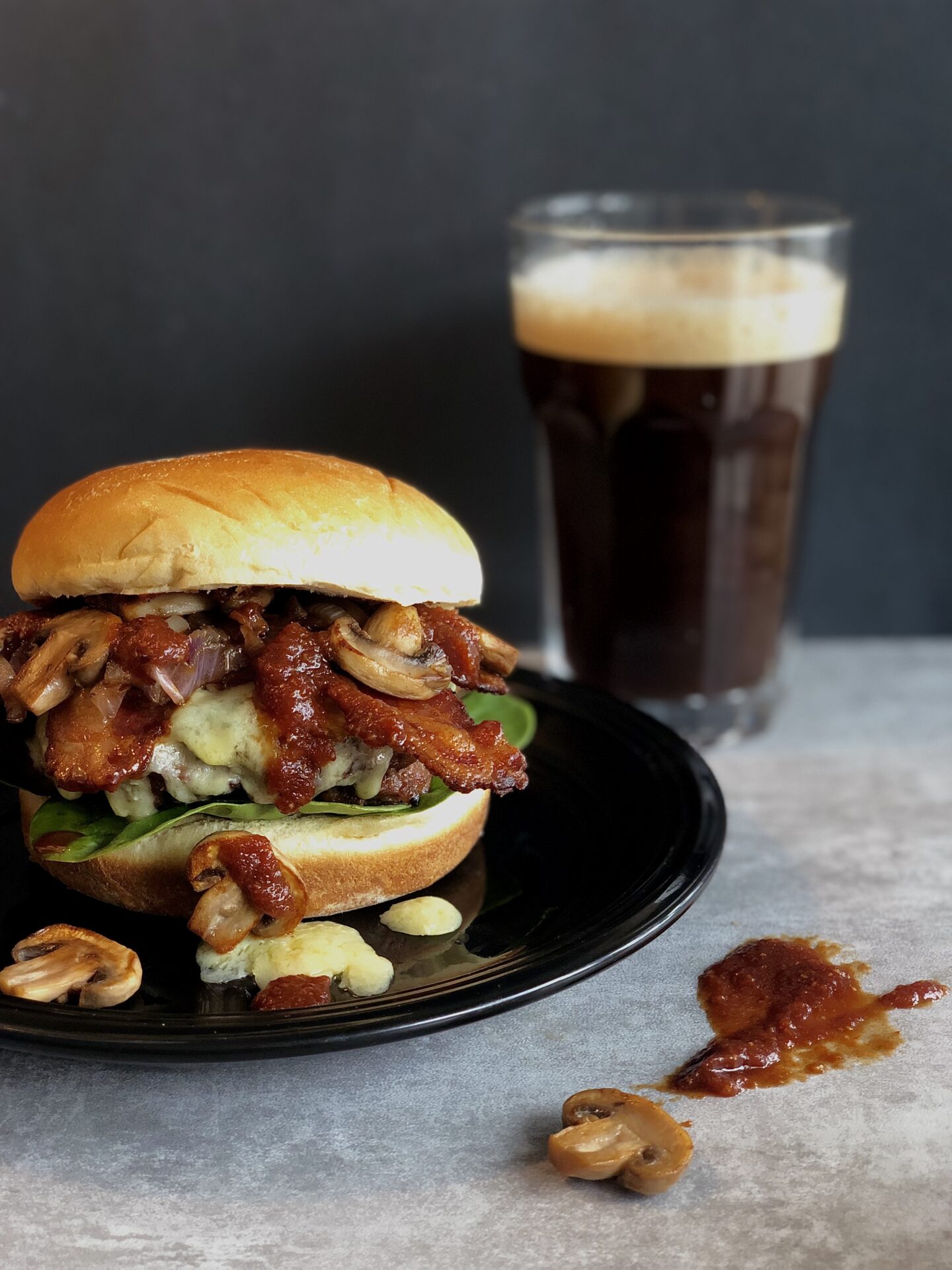 A Guinness Bacon Burger with Mushrooms and Irish Cheddar is seen on the left side of the photo. A pint of Guinness beer and a drip of homemade Guinness barbecue sauce can be seen on the right.