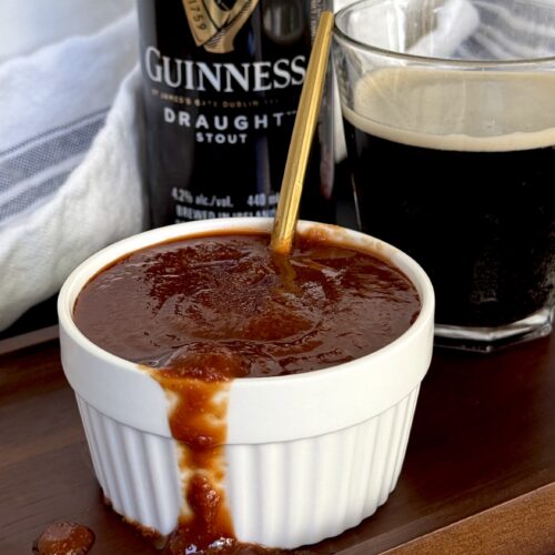 A bowl of Guiness Barbecue Sauce overflows onto a wooden board. A can and glass of Guiness beer can be seen in the background.