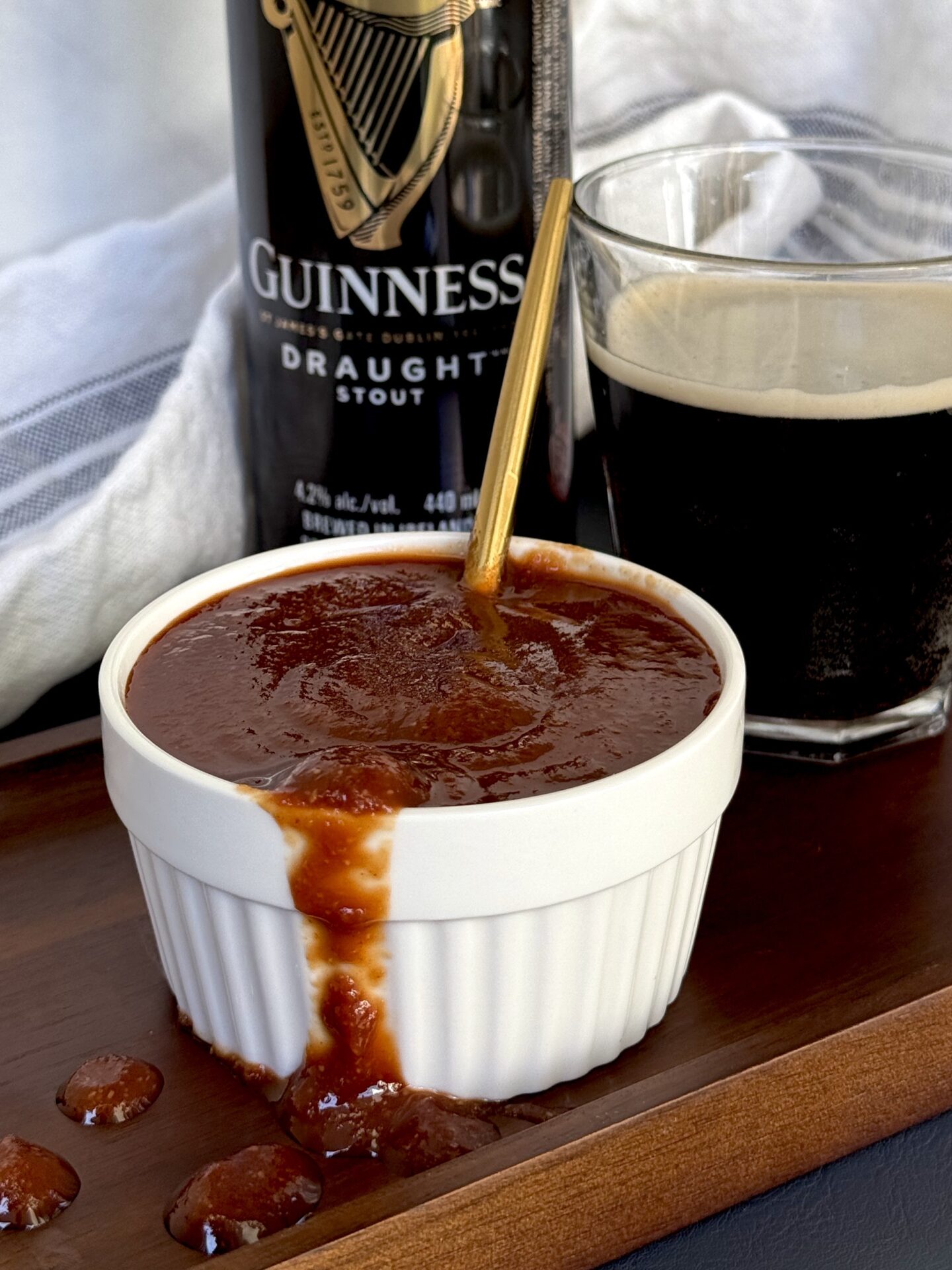 A bowl of Guiness Barbecue Sauce overflows onto a wooden board. A can and glass of Guiness beer can be seen in the background.