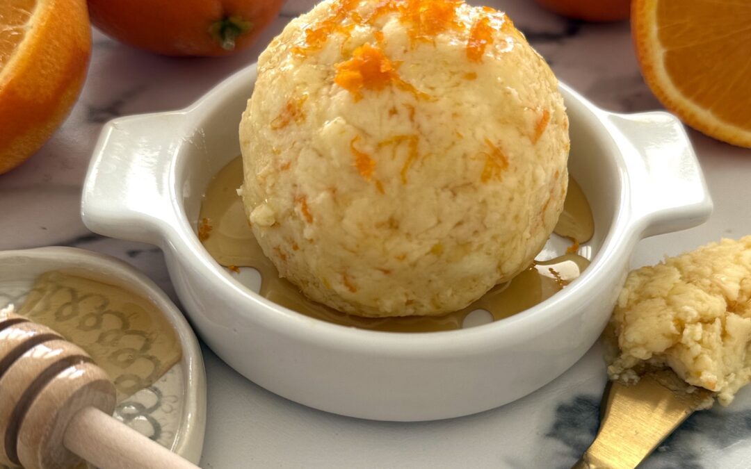 Orange and Honey Butter is shaped into a ball, placed in a shallow butter dish, drizzled with extra honey and garnished with fresh orange zest. Fresh oranges and a honey dipper complete the image.