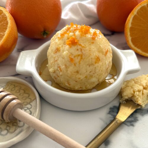 Orange and Honey Butter is shaped into a ball, placed in a shallow butter dish, drizzled with extra honey and garnished with fresh orange zest. Fresh oranges and a honey dipper complete the image.