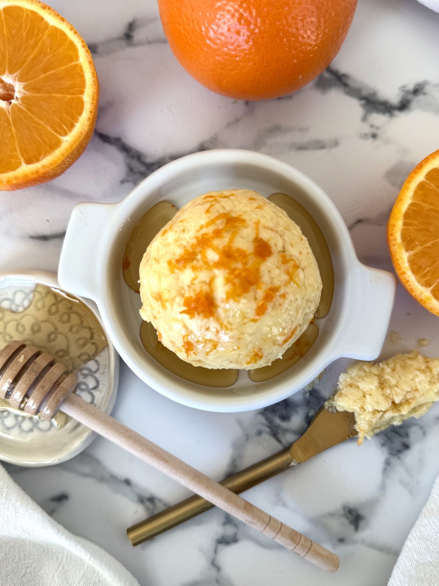 A sphere of Orange and Honey Butter is seen from above. drizzled with honey and sprinkled with fresh orange zest.   The butter rests in a dish on a white marble counter, surrounded by fresh oranges, a butter spreader and a honey dipper.