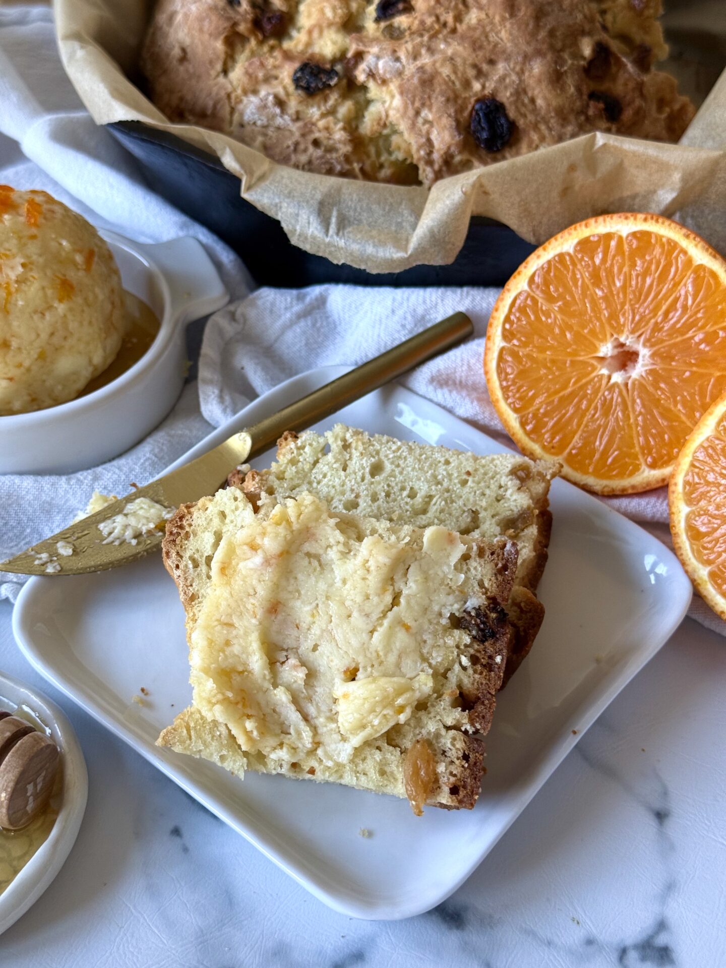 Homemade Orange and Honey Butter is slathered on slices of freshly baked Irish soda bread.  A loaf of the soda bread can be seen in a cast iron skillet in the background.  Fresh oranges and a honey dipper complete the image.  