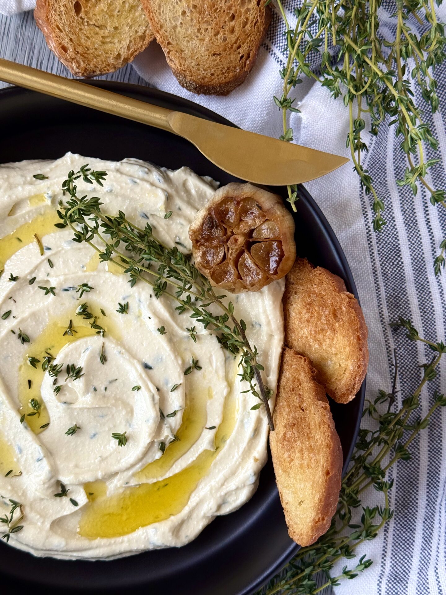 IMG_5606 A black bowl filled with swirled Whipped Feta with Roasted Garlic and Thyme dip is seen on a grey wood table. The dip is garnished with drizzles of olive oil, a head of roasted garlic and sprigs of fresh thyme. The dish is surrounded by slices of toasted baguette and sprigs of fresh thyme. A gold spreader sits on the edge of the dish.