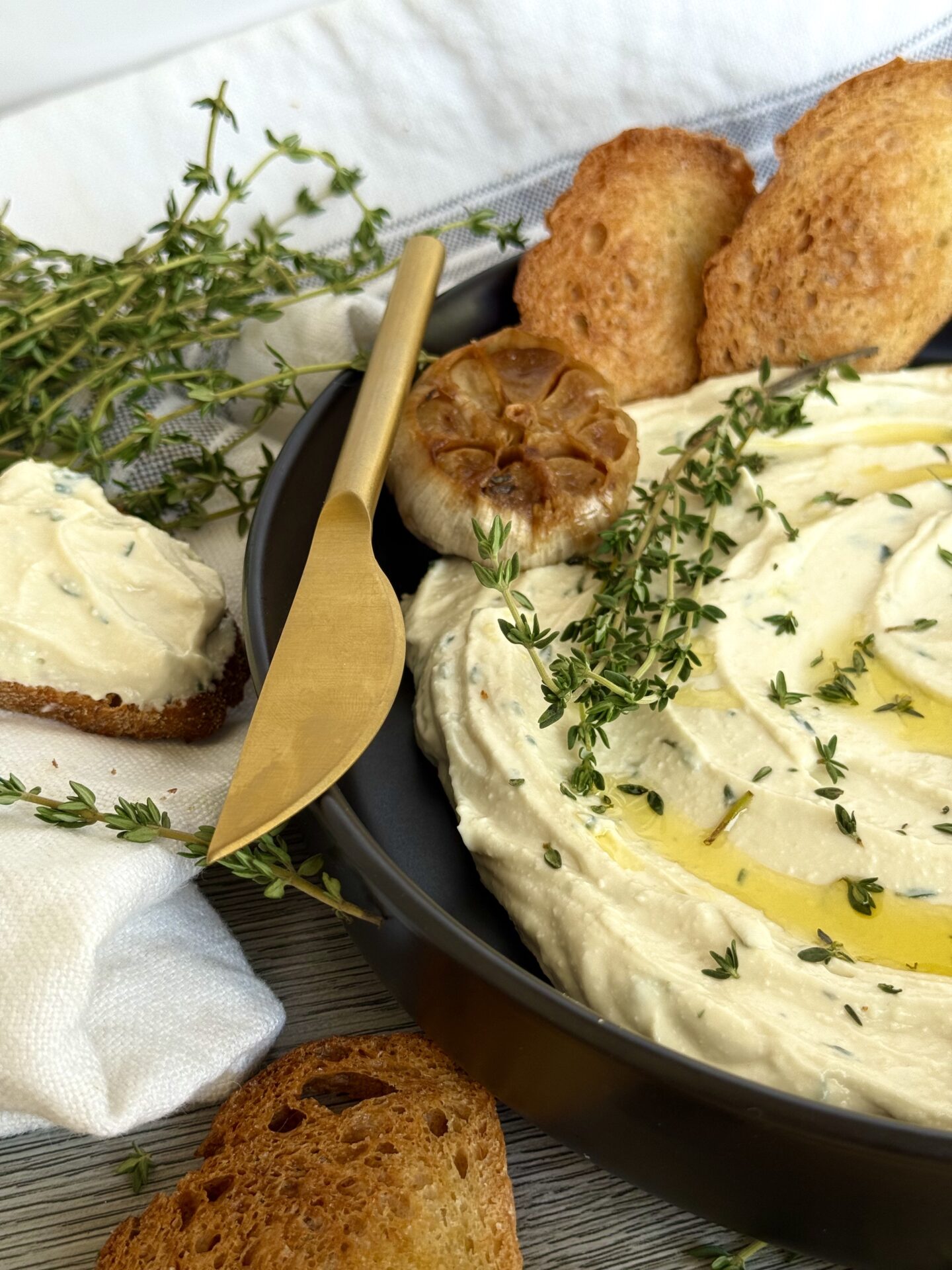 IMG_5621 Half of a black dish filled with Whipped Feta with Roasted Garlic and Thyme dip fills the right side of the image. The dip is lavishly garnished with olive oil and fresh thyme. A white and grey striped kitchen towel surrounds the bowl and sprigs of fresh thyme and toasted baguette slices complete the image.
