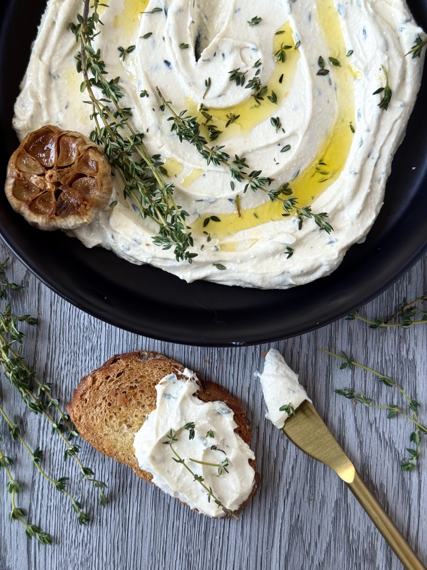 IMG_5624 A black dish is seen from above, filled with swirled Whipped Feta with Roasted Garlic and Thyme. In front of the bowl sits a slice of toasted baguette slathered in the dip. A gold spreader, a head of roasted garlic and sprigs of fresh thyme complete the picture.